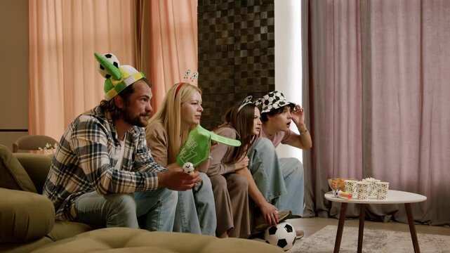 Group of friends wearing festive hats and holding soccer-themed props watch a game together on a comfortable sofa in a cozy living room setting