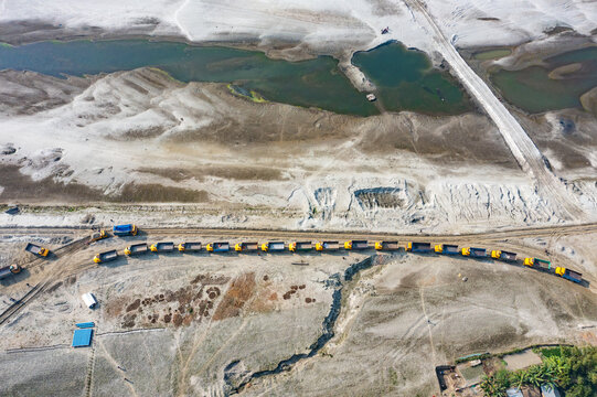 Aerial view of a long line of yellow dump trucks parked on a dirt road along the sandy banks of the Jamuna River Char in Tangail District, Dhaka Division, Bangladesh.