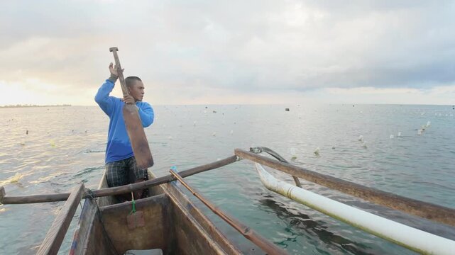 An adult indonesian seaweed farmer rowing with a paddle on a traditional wooden boat on aquaculture farm at sea in indonesia, southeast asia
