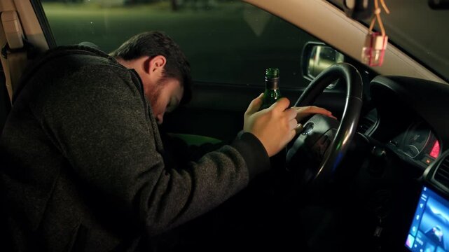 Drunk man holds green bottle with whiskey inside vehicle and leans hands on steering wheel. Vehicle setting at night with asleep caucasian man clutching bottle. Driver under the influence of alcohol