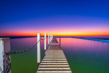 Fototapeta premium Leading lines from the timber swimming platform guide the eye across North Narrabeen Rockpool at sunrise, featuring calm water, pastel skies and Sydney coastal scenery.