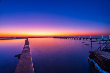 Fototapeta premium Timber swimming platform extending across North Narrabeen Rockpool during sunrise on Sydney's Northern Beaches, captured with smooth long exposure water and vibrant colours.