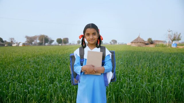 Well-dressed village school girl smiling while standing in her agricultural field. Medium shot of an Indian kid in a school uniform carrying her books and a school bag - girl child education