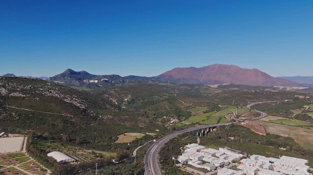 Aerial view of a stone quarry with a processing plant and asphalt plant near a highway viaduct and green hills under a clear blue sky in Manilva, Andalusia, Spain.