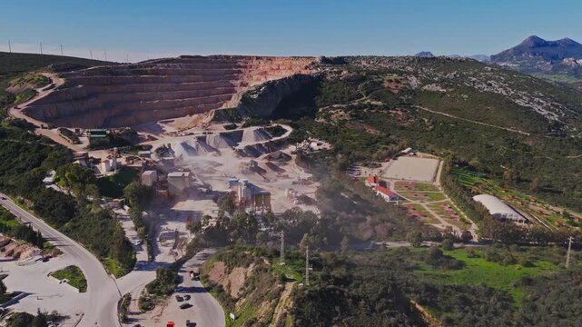 Aerial view of a large stone quarry with a processing plant and asphalt plant nestled among green hills under a clear blue sky in Manilva, Andalusia, Spain.