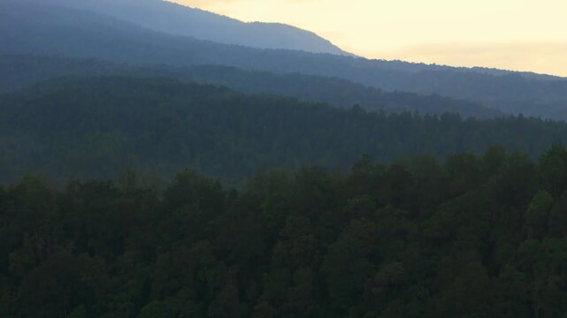 Aerial view of lush forested hills and layered mountain ridges under a hazy sky at dawn in Sukabumi, West Java, Indonesia.