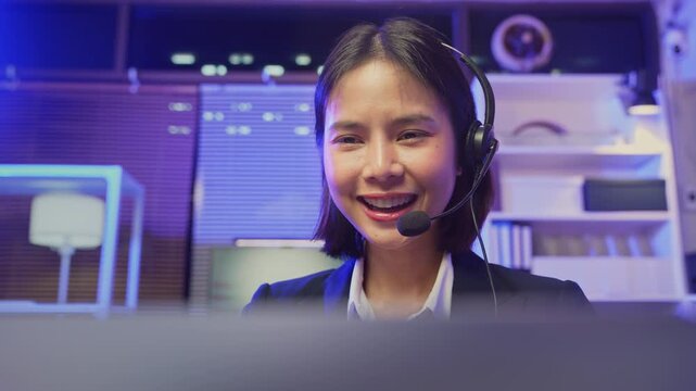 Smiling young Asian customer service representative wearing microphone headset and working on laptop in modern office.