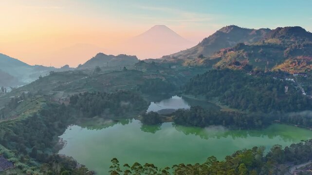 Aerial view of a serene lake covered in morning mist surrounded by lush green hills and a distant volcanic peak during golden hour in Pekalongan, Central Java, Indonesia.