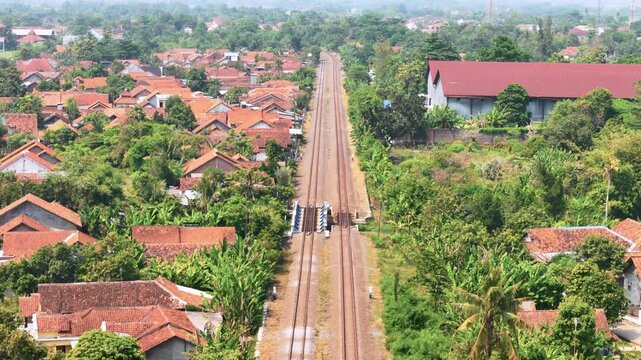 Aerial view of a long railway track stretching through a residential neighborhood with orange rooftops and dense green trees in Pekalongan, Central Java, Indonesia.