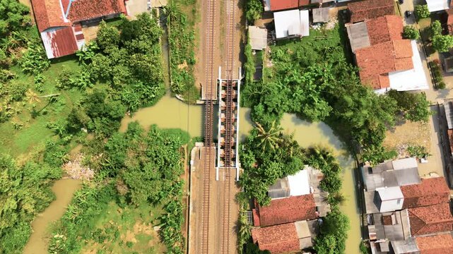Aerial view of a railway bridge crossing a narrow river surrounded by red-tiled houses and lush green vegetation in Pekalongan, Central Java, Indonesia.
