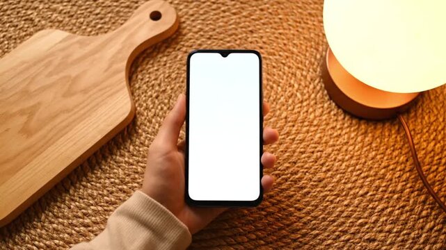 top down view of a hand holding a smartphone with a blank white screen on a woven mat next to a cutting board and lamp