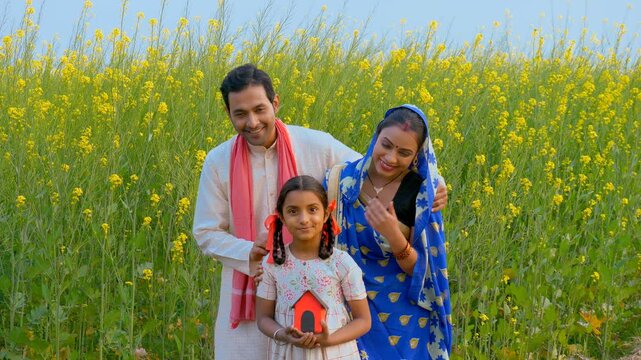 Indian village family standing against the Sarso field with a dream house model. Medium shot of a village couple desiring a new house while their daughter is holding a model hut - new home