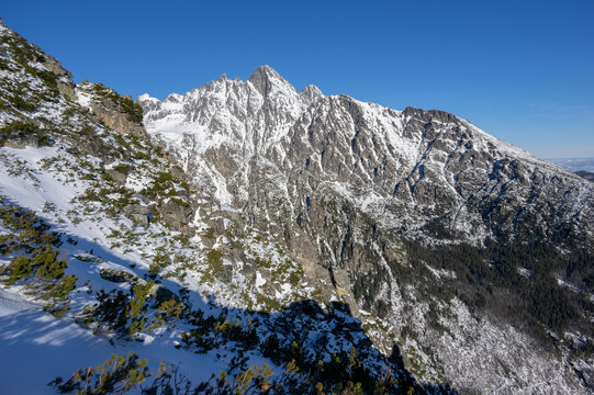 View of Slavkovsky nos peak with snow-covered rocky slopes and green shrubs under a clear blue sky in Vysoke Tatry, Presov Region, Slovakia.