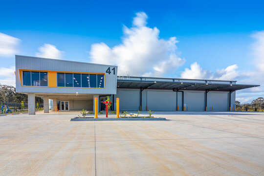 Modern industrial warehouse and office facility with multiple roller doors, loading area and expansive concrete hardstand under a blue sky. Commercial logistics and distribution building in Australia.