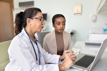 Naklejka na ściany i meble Senior female doctor giving consultation to young African patient, explaining medical records, expertise results, examination report, pointing at laptop at workplace, speaking to woman