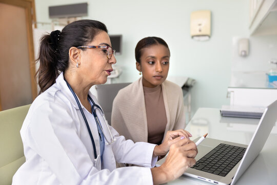 Senior female doctor giving consultation to young African patient, explaining medical records, expertise results, examination report, pointing at laptop at workplace, speaking to woman