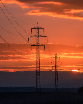 View of high voltage power lines and electricity pylons silhouetted against a vibrant orange sunset sky with scattered clouds Kyjov, South Moravian Region, Czechia.