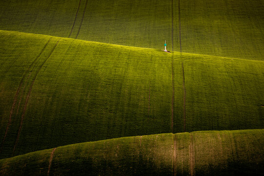 View of rolling green agricultural hills with tractor tracks and a small marker on a ridge in Cejkovice, South Moravian Region, Czechia.