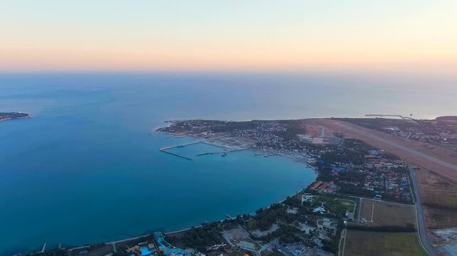 Gelendzhik, Russia. Aerial panorama over the horseshoe-shaped bay, Thin Cape and airport runway silhouetted against a soft sunset sky, turquoise Black Sea waters stretching toward the Thick Cape. Dro