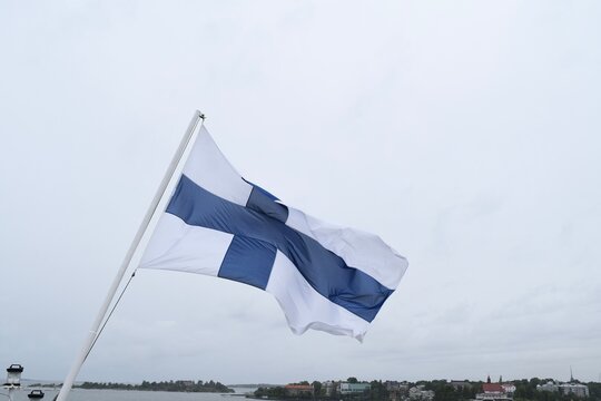 National flag of Finland waving on a flagpole against a cloudy sky with Helsinki coast in the background 