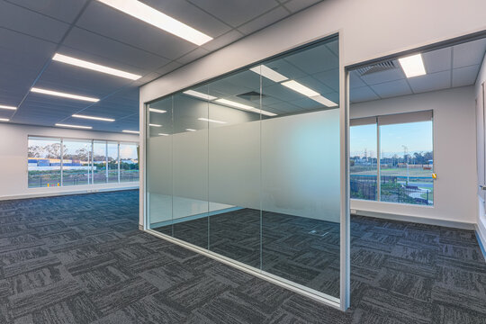 Modern office interior with glass partition meeting room, open-plan workspace, carpet tile flooring and large windows overlooking a commercial estate.