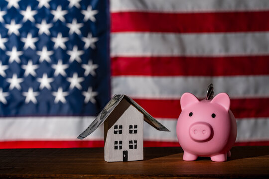 Piggy bank and toy house with dollar roof on US flag background. Pink piggy bank with coins next to a small wooden house with dollar bill roof against the USA flag