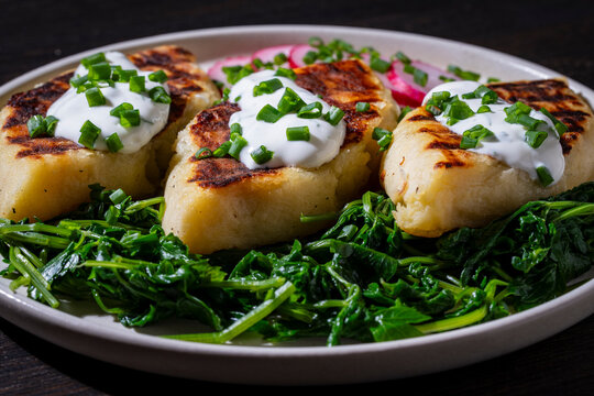 Potato zrazy cutlets with sour cream and spinach on plate. Fried potato cakes or zrazy served with white sauce green spinach and radish on ceramic plate closeup