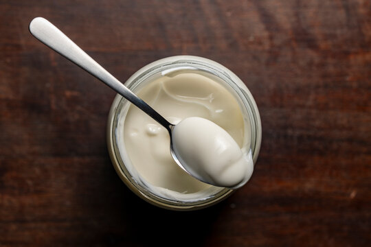 Fresh organic yogurt in a glass jar with a spoon on wooden background. Top view of creamy white yogurt or sour cream inside a glass jar with metal spoon on dark wooden table