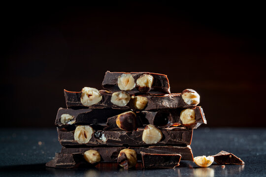 Stack of broken dark chocolate bars with whole hazelnuts. Closeup of dark chocolate pieces with whole hazelnuts stacked on a black background