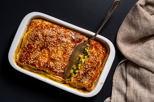 Homemade greens and egg pie with sesame seeds in baking dish. Top view of savory spinach and egg pastry pie with sesame seeds in white ceramic baking pan on black background