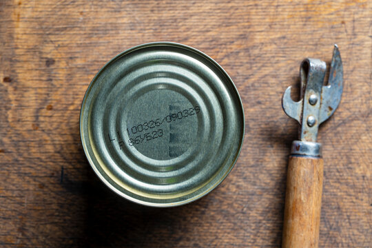 Closed tin can with expiration date and vintage opener on wooden table. Top view of a sealed metal tin can with printed codes next to a retro can opener on rustic wooden background