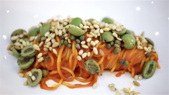 Plate of gourmet spaghetti with tomato sauce topped with green olives and pine nuts served in white restaurant dish.