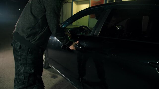 Drunk adult man with green bottle in grey cloth opens the unlocked door of black car at night on empty road. Male hand opens vehicle door in darkness in city. The danger of drunk driving. No face
