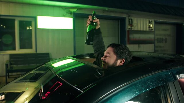 Excited bearded caucasian man celebrates with opened sunroof of car during night in city. Vibrant neon scene of joyful happy driver raising green bottle with whiskey. Drinking alcohol while driving