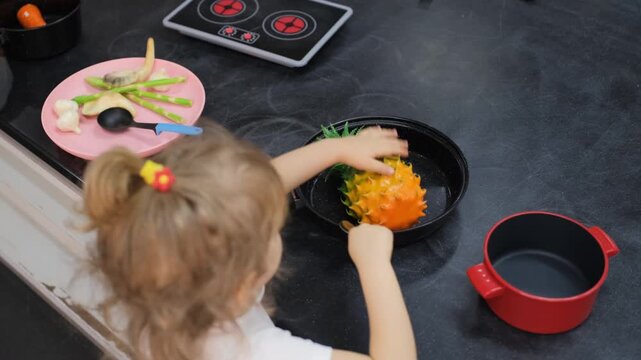 Cute little girl pretending to fry a toy pineapple in a toy pan on a play kitchen stove in an indoor play area. Imaginative play concept
