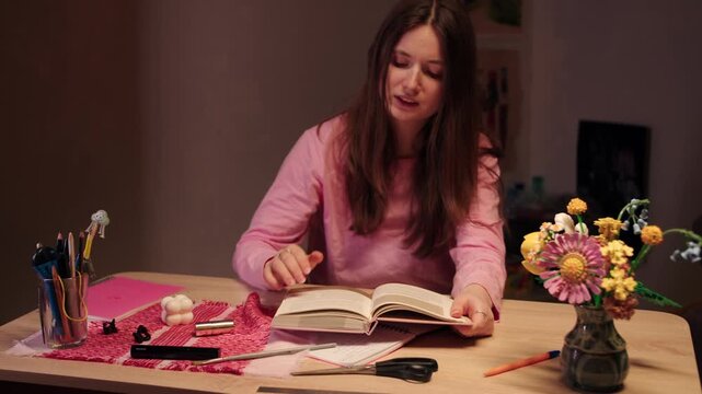 A tired beautiful young caucasian woman in a pink sweater sits at a table surrounded by stationery and a bouquet of flowers, yawns and reads a book. Boredom and procrastination during homeschooling