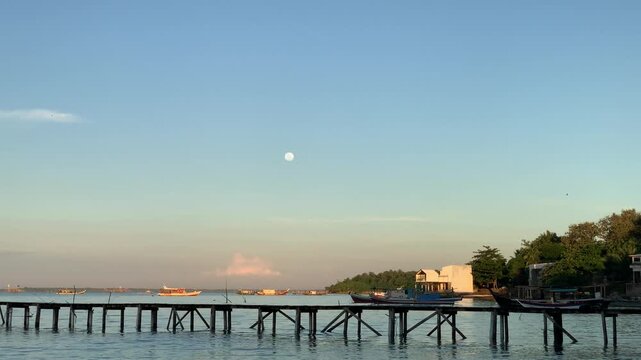 4K scenic view of Tanjung Lesung Beach, Banten, Indonesia with wooden pier, fishing boats, calm sea, and visible moon above the horizon. Ideal for travel, tourism, coastal, lifestyle, and nature theme