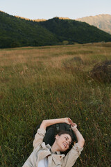 Naklejka premium Young woman with short brown hair lies on tall grass in a natural field surrounded by hills, enjoying a peaceful moment with a relaxed and content expression.