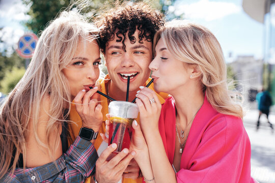 Young three stylish friends posing in the street. Fashion man and two cute female dressed in casual summer clothes. Smiling models having fun. Cheerful women and guy outdoors, Hold and drink lemonade
