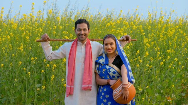 Handsome farmer and his wife in traditional dresses standing in their mustard  or sarso field. Medium shot of an Indian traditional married couple smiling cheerfully at each other - farmer's family