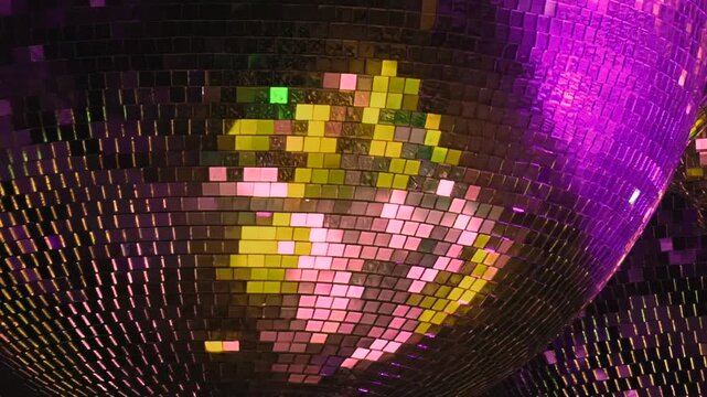 A dazzling disco ball, aglow with dynamic purple, green, and gold light, rotates slowly. Its mirrored facets scatter brilliant, ever-changing patterns across a festive dance space at night.