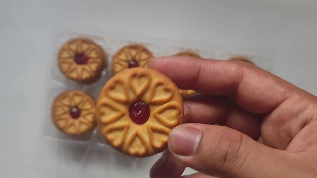 Macro footage of a hand picking up a sweet jam-filled cookie from a plastic tray and presenting it close to the camera on a white background