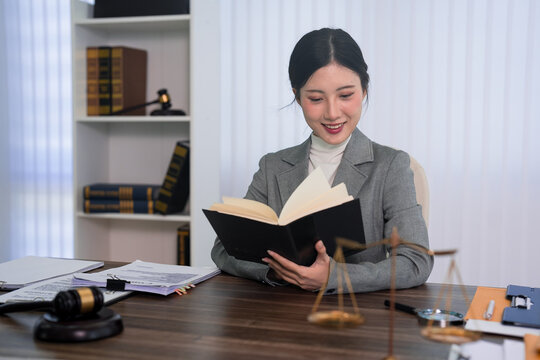 young Asian lawyer woman reading law book studying legal document case research working at professional law office desk with gavel justice scale bookshelf