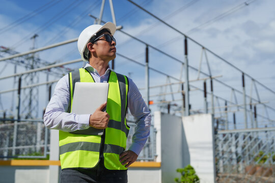 Asian male engineer in hard hat and safety vest holding a laptop, standing at an electrical power substation with high voltage equipment visible and blue sky