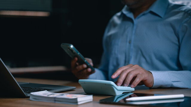 Professional man sits wooden desk inside dark office operating calculator smartphone near laptop stack cash digital tablet managing personal finances nighttime alone working home late evening indoor.