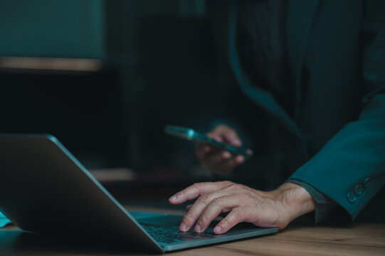 Professional businessman wearing dark suit works late night inside dim office environment, typing laptop keyboard keys holding smartphone over wooden desk surface performing overtime shift digital.