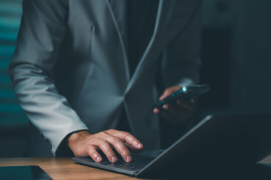 Unrecognizable businessman wearing gray suit types laptop keyboard holding smartphone wooden desk dimly lit office setting late night work session digital business project professional corporate.