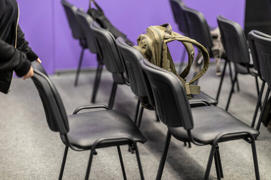 A person arranges rows of empty black chairs in an indoor conference or event space, with a militarian backpack resting on one of the seats in the foreground, against a distinct purple wall surface