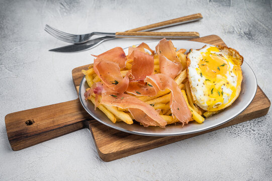 Huevos rotos con jamon with fried potatoes and runny egg served on oval plate top view on light stone background