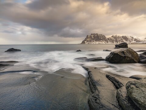 Smoothly polished rocks on Utakleiv beach in a dramatic cloudy atmosphere, snow-capped mountains in the background, Vestvagoy Island, Lofoten, Norway